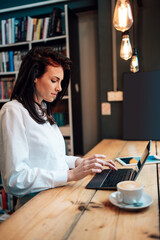 Beautiful female student or young business woman using her laptop computer and standing next to the window in a cozy working cafe bar.