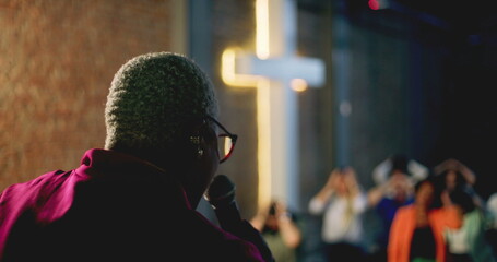 Woman holding the Bible and addressing the congregation from a stage with a glowing cross in the...
