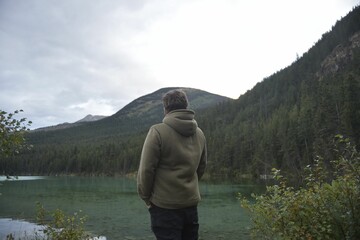 Fototapeta premium Man in a hoodie stands at the edge of a serene alpine lake.