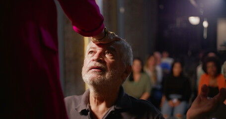 Elderly man kneeling during church blessing, hand placed on his forehead by spiritual leader, expression of faith, devotion, and humility, indoor religious gathering