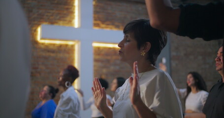 Woman praying with closed eyes and raised hands in a diverse group worship setting, illuminated...