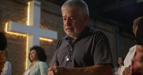 Close-up of elderly man reflecting during prayer in church, hands clasped in devotion, illuminated...