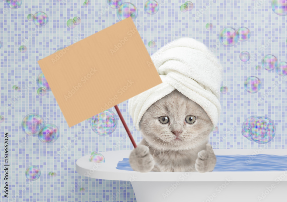 Poster cute kitten with towel on it head takes the bath at home, showing blank placard and thumbs up gestur