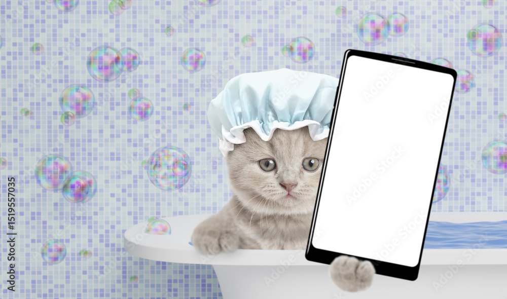 Poster cute kitten wearing shower cap takes the bath at home and shows big smartphone with white blank scre