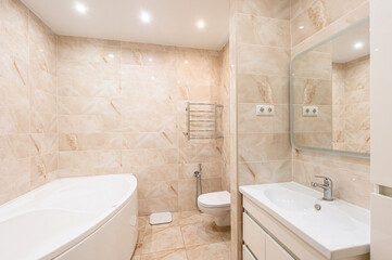 bright bathroom with beige marble tile, a white tub, toilet, and sink, and a mirrored cabinet....