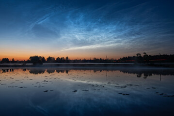 A calm lake at dusk with a misty pier, calm reflections, tree silhouettes and bright, noctilucent clouds in a colorful sky, silver clouds