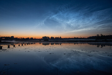 A calm lake at dusk with a misty pier, calm reflections, tree silhouettes and bright, noctilucent clouds in a colorful sky, silver clouds
