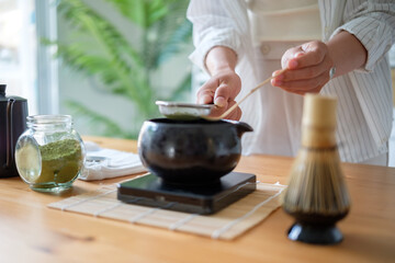Close up of hands sifting matcha powder into a traditional black bowl for tea preparation at home.