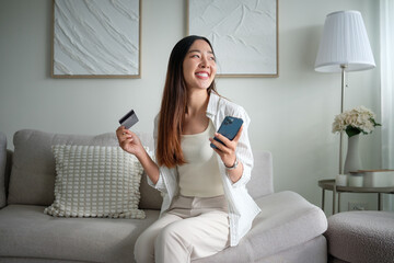 Casual asian woman sitting on sofa enjoying digital banking or mobile purchase on her smartphone.