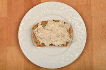 A top-down shot features a piece of bread generously topped with a creamy tuna spread, centrally placed on a white patterned plate atop a wooden surface