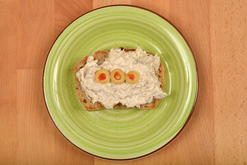 A top-down shot features a piece of bread topped with a creamy tuna spread and garnished with three green olives, centrally placed on a light green spiral-patterned plate on a wooden surface