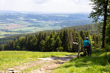 Fototapeta premium A man in a helmet on a mountain bike is observing a cycling trail on green grass with a view of the valley and the forest.