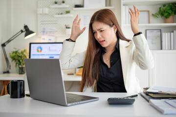 Frustrated Asian businesswoman raises hands in anger while staring at laptop screen in modern office environment.