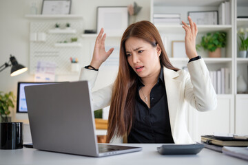 Young Asian businesswoman in white blazer raises hands in frustration while looking at laptop screen during office work.