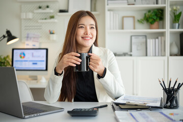 Smiling young Asian businesswoman holding a black coffee mug while taking a break in a bright modern office.