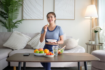 Young woman in activewear enjoying a nutritious breakfast in a cozy living room setting with fresh fruits and salad on the table.