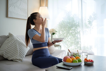 Young woman in sportswear relaxing on the sofa and enjoying a nutritious meal with a plate of fresh fruits and salad nearby.