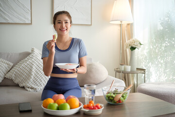 Cheerful woman in activewear holding a bowl and eating a healthy snack at home, surrounded by natural light and nutritious food.