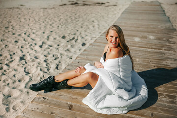 A serene beach moment with a woman wrapped in white blanket enjoying the sun on a wooden dock by the sea at midday