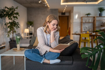 Obraz premium Woman reading a book at home on the sofa in her living room