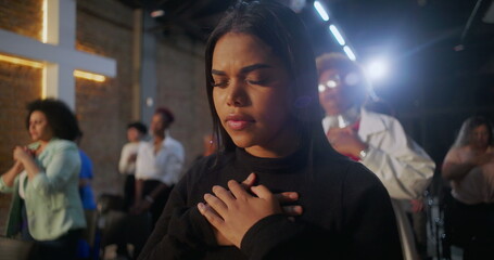 Fototapeta premium Women in prayer during church service, hands on chest in devotion, illuminated cross in the background, community gathering in spiritual reflection and worship