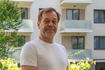 Portrait of a smiling man 50-55 years old in a white T-shirt on the background of a residential building with balconies.