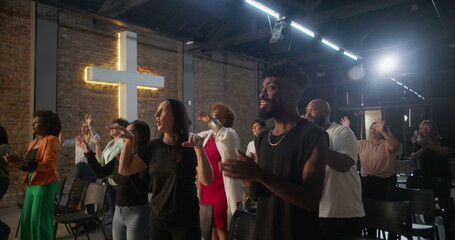 Multicultural congregation clapping and smiling during church worship, illuminated cross in...
