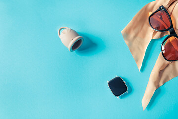 Modern flat lay of menstrual cup, smart watch, sunglasses and scarf on bright blue background....