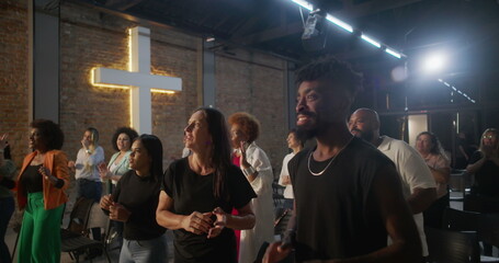 Multicultural congregation clapping and smiling during church worship, illuminated cross in...
