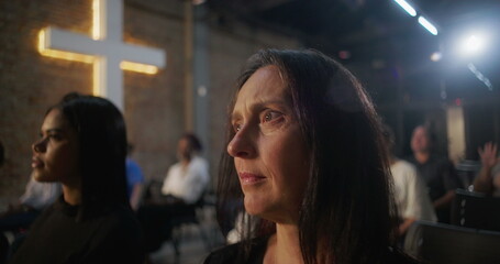 Woman sitting in church with thoughtful expression, illuminated cross and congregation in background, reflecting during worship service in sacred community gathering