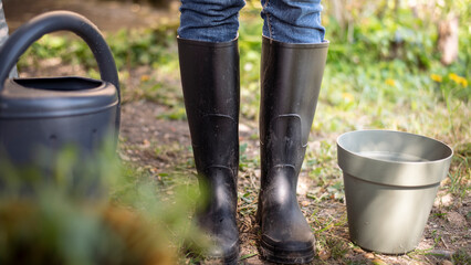 Close-up view of a person wearing black rubber gardening boots standing next to a flower pot and a watering can in an outdoor garden. The image evokes a peaceful gardening moment, with natural light.