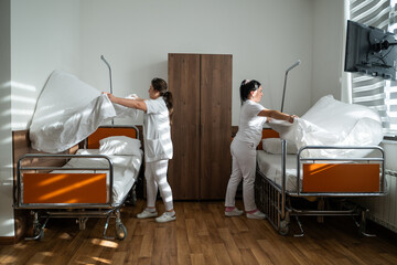 Two nurses in uniforms cleaning and organizing a hospital room by changing the bed linens and...