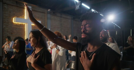 African American man in emotional worship, hand raised and over heart, glowing cross in background,...