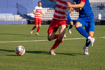 Two soccer players in red and blue uniforms competing for the ball during a match on a green artificial turf field.