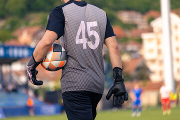 Back view of a goalkeeper holding a football while walking across the field, wearing gloves and a jersey with the number 45.