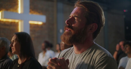 Man bowing head in prayer, hands clasped, deeply engaged in spiritual practice in a church, illuminated cross on a brick wall in the background