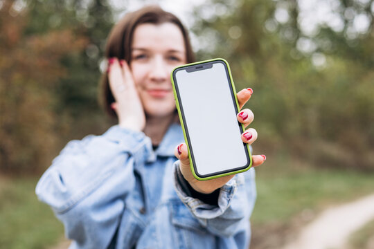 Smiling young woman showing blank smartphone screen to camera outdoors.