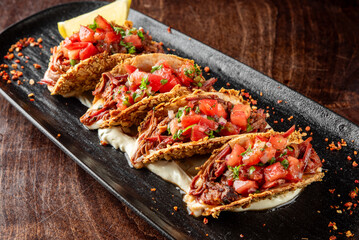 Four crispy tacos with shredded meat, diced tomatoes, onions, and cilantro on a black plate with a lime wedge. Garnished with chili flakes, served on a wooden table