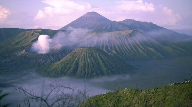 Beautiful view of Mount Bromo with smoking crater, green hills and morning mist covering the valley.