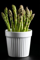 Fresh green asparagus spears standing upright in a white ceramic cup against a black background