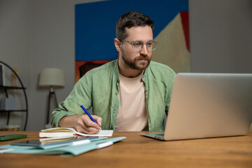 Bearded man reviewing notes on desk while working online with laptop and papers