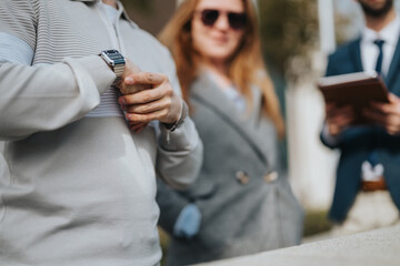 A focused image of a person's hand wearing a smart watch, with blurred individuals in professional attire indicating a formal meeting or business environment in the background.