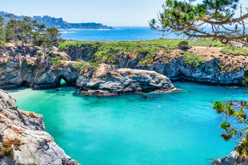 Breathtaking view of China Cove at Point Lobos State Natural Reserve, California. Turquoise waters, rocky cliffs, and vibrant coastal vegetation under a clear summer sky