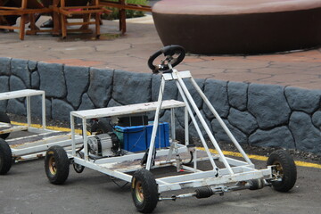 A simple homemade go-kart with a visible engine and frame is parked on a paved area near a stone wall, showcasing a DIY vehicle project or recreational engineering build.