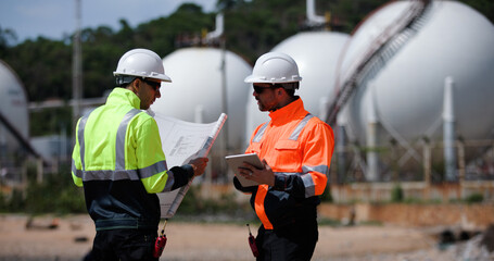 Two man industrial engineer with green and orange safety suit working with blueprint drawing at Refinery and gas petrochemical plant