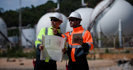 Two man industrial engineer with green and orange safety suit working with blueprint drawing at Refinery and gas petrochemical plant