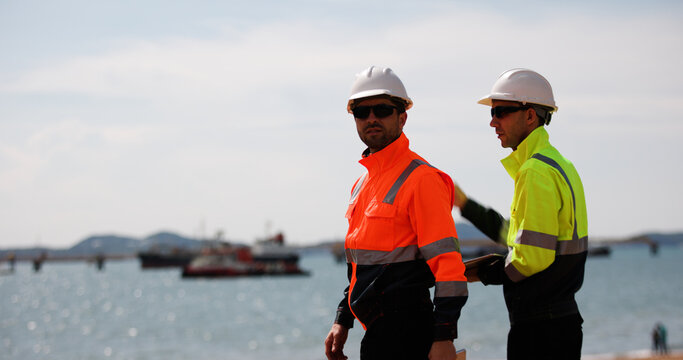 Two male power plant engineers work with a blueprint and a digital tablet by the seaside at a refinery and gas petrochemical plant - Powered by Adobe