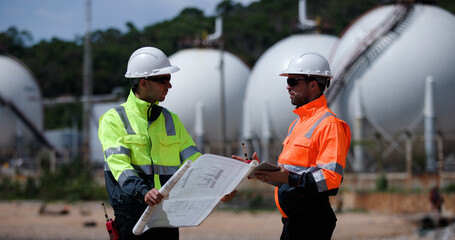Two man industrial engineer with green and orange safety suit working with blueprint drawing at Refinery and gas petrochemical plant