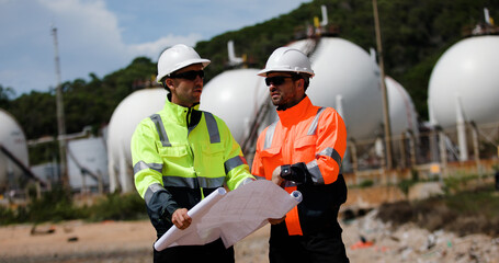 Two man industrial engineer with green and orange safety suit working with blueprint drawing at Refinery and gas petrochemical plant