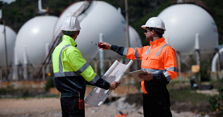 Two man industrial engineer with green and orange safety suit working with blueprint drawing at Refinery and gas petrochemical plant